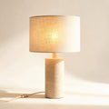 Lampe à poser Trocadéro, pied colonne en travertin et abat-jour en lin, allumée, sur fond crème