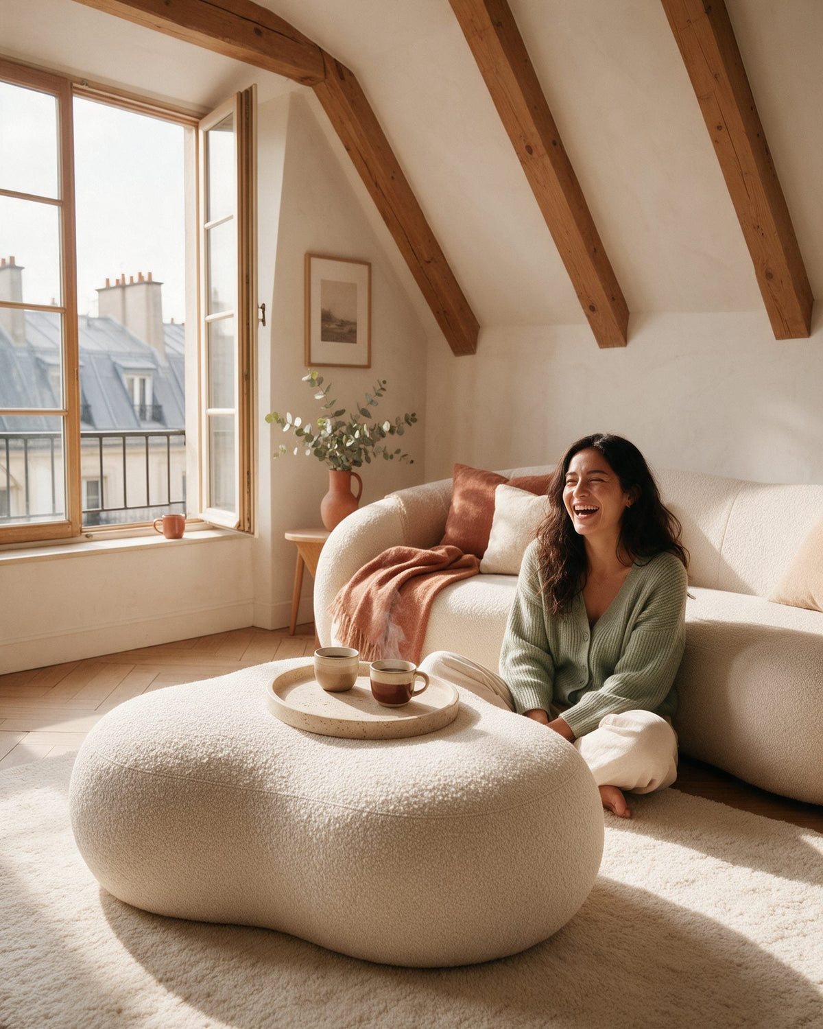 Pouf Nuage en bouclette écrue devant un canapé arrondi, avec un plateau et deux tasses de café ; une femme rit, assise sur le tapis