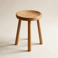 Tabouret Contrescarpe en chêne massif huilé, assise ronde légèrement creusée et trois pieds fuselés, sur fond crème