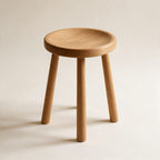 Tabouret Contrescarpe en chêne massif huilé, assise ronde légèrement creusée et trois pieds fuselés, sur fond crème