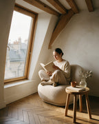 Une femme lit dans un fauteuil en bouclette sous les toits de Paris ; à côté, le tabouret Contrescarpe en chêne porte un livre, une tasse et un petit vase
